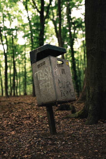 A weathered grey metal mailbox with graffiti markings and a black open lid, mounted on a tilting post in a woodland setting. The mailbox is situated on a dirt path covered with fallen leaves and small twigs, surrounded by tall trees with coarse bark and green foliage, which filter diffused sunlight and create a dappled light effect on the ground. The mailbox appears aged, with visible rust, scratches, and graffiti tags in black ink on its surface. It is positioned slightly off-center to the left, with the trees in the background extending into the distance, giving a sense of depth. The natural environment and the presence of the mailbox suggest it is in an outdoor area that may be accessible for private or independent waste collection, aligning with the concept of alternative waste handling methods. The overall scene is calm and quiet, emphasizing a typical woodland backdrop with elements that mirror the need for rubbish removal services in rural or semi-rural locations.
