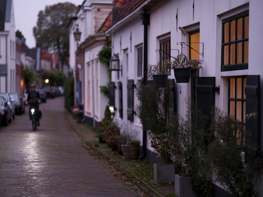 A narrow residential street in a town during dusk, featuring a row of traditional white-painted cottages with black window frames and shutters. The cottages have small black wall-mounted lantern lights and window boxes filled with pink and purple flowers. Potted plants and shrubs are situated along the sidewalk in front of the houses. The cobblestone pavement reflects the soft lighting, creating a calm atmosphere. In the background, a cyclist is riding towards the camera, and parked cars line the street on the left side. The scene captures the quiet charm of a historic neighbourhood, with the subtle evening light highlighting the textures of the buildings and street. This setting illustrates the local environment where private and independent rubbish removal services, such as those offered by Rubbish Removal Holland Park, may operate in similar residential areas, supporting efficient waste management outside of local authority collection schedules.
