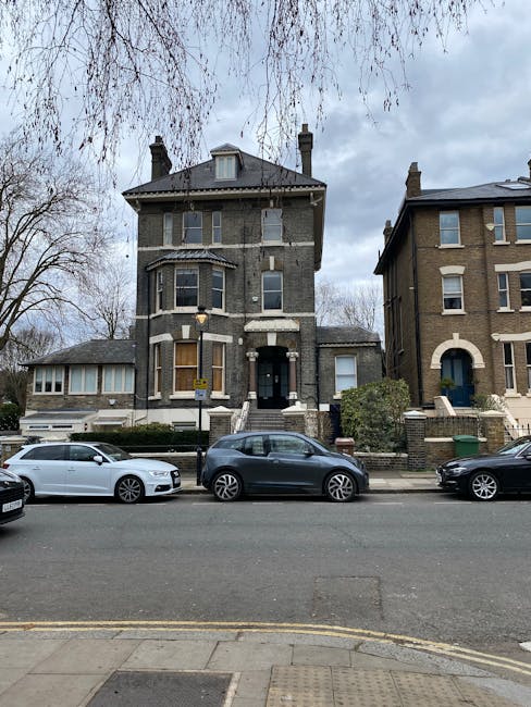 A tall, multi-story Victorian-style house constructed of dark grey brick with contrasting white trim around the windows and door. The building features a prominent central bay window on the second floor and a small, pitched roof above the entrance, which is accessed via a short staircase with black railings. The house is situated on a residential street with parked cars, including a white sedan, a dark grey hatchback, and a black vehicle, lined up along the curb in front. To the left of the main building, there is a single-story extension with large windows, possibly a conservatory or porch. On the right side, part of another similar Victorian-style house with a light brown brick facade and arched doorway is visible. Trees with sparse, leafless branches frame the scene, indicating a late autumn or winter setting. The sky is cloudy, and the environment appears calm and residential. This scene exemplifies private property and on-site waste management typical of independent rubbish collection services like Rubbish Removal Holland Park, contrasting with council-led refuse collection.