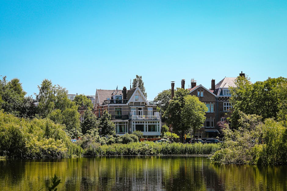 A residential scene depicting a row of multi-story houses with pitched roofs and brick facades, situated behind a lush, green riverside area. The houses feature large windows, some with balconies, and vary in architectural details and finishes such as white trims and dark brickwork. Surrounding the houses are tall deciduous trees with dense foliage, creating a natural border along the riverbank. In the foreground, calm water reflects the greenery and buildings, while the sky above is clear with a vibrant blue hue, indicating a bright, sunny day. The environment appears peaceful and well-maintained, with the natural landscape integrated into the urban setting, reflecting typical outdoor spaces associated with private residential properties. This scene emphasizes the importance of managing outdoor waste and debris resulting from regular residential maintenance, aligning with professional rubbish removal and waste management services in the area.