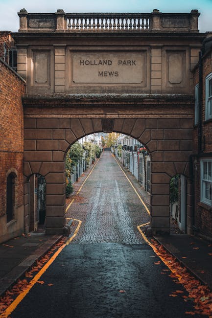 A narrow, slightly inclined cobblestone street extending through an arched gateway constructed from weathered sandstone blocks, with the inscription 'HOLLAND PARK MEWS' engraved on the upper section. The street is flanked by wrought iron fences and residential buildings with brick facades, and the ground is scattered with fallen leaves. The scene is captured during daylight hours with overcast sky, creating diffuse natural lighting that highlights the textures of the stone and the subtle color variations in the brickwork. This setting, typical of an urban residential area, suggests an environment where private waste collection or on-site clearance services, such as those offered by Rubbish Removal Holland Park, might be relevant for maintaining cleanliness and managing rubbish outside of regular municipal collection. The environment emphasizes the context of discreet, local rubbish removal in a historic, well-maintained neighbourhood, where independent waste handling could occur near the properties lining the street.