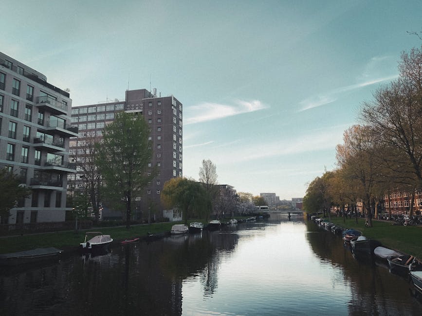 A canal running through an urban area during daytime, with modern multi-storey residential buildings with balconies on the left side and a row of trees with green and brown foliage on the right side. Along the edges of the canal, various small boats are moored, with some partially covered and others exposed to the water. The water in the canal is calm, reflecting the sky and surrounding structures, while a bridge is visible in the distance spanning the canal. The sky has a pale blue tone with high, wispy clouds, and the lighting indicates a bright, clear day. This scene captures a peaceful urban environment, where private boat moorings and residential buildings are set close to nature, aligning with the concept of alternative waste management or on-site disposal options that Rubbish Removal Holland Park might facilitate in such city settings.