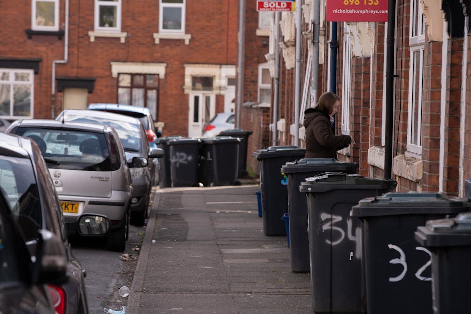 A residential street scene showing a row of large black wheelie bins lined up along the sidewalk in front of brick terrace houses. The bins, used for waste disposal, are positioned close together and feature white painted numbers on their sides. A woman dressed in a brown coat stands near one of the bins, appearing to be in the process of disposing of rubbish or checking the contents. Several parked cars are visible on the street, with a variety of vehicles including a silver hatchback nearest the camera and other cars further down the road. The environment is typical of an urban residential area, with cobbled pavement, brick facades, and framed windows. Some of the houses have small front gardens or steps leading to entrances, and there is a red street sign with white text attached to one of the buildings. The scene is captured in natural daylight, with even lighting and no visible shadows, indicating an overcast or partly cloudy day. This image illustrates a typical setting for private waste collection or rubbish removal services in an urban environment, with a focus on the collection points for the black waste bins commonly used in local waste management or alternative disposal arrangements.