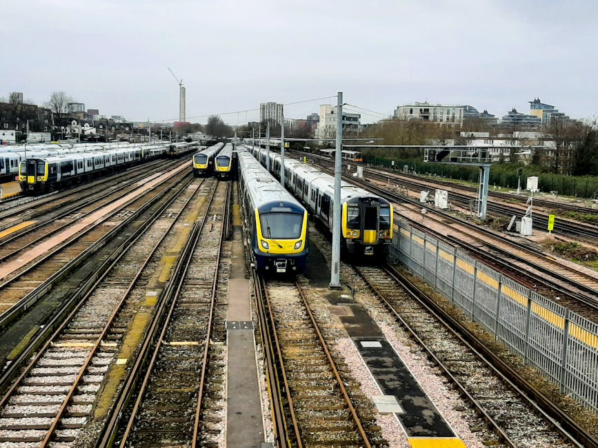 A train depot with multiple sets of railway tracks occupied by several modern trains. Three trains are positioned side by side in the foreground, with the central train front and center, featuring a streamlined, aerodynamic design and painted in yellow and dark grey. To the right, a black train is partially visible, coupled to a grey and yellow barrier fence that runs parallel to the track. In the background, additional train carriages are parked on neighboring tracks, with some vehicles also visible on a distant road. Overhead electric lines and supporting pylons stretch across the scene, indicating an electrified rail system. The surrounding environment consists of urban elements, including multi-storey buildings, trees, and a cloudy sky, suggesting an overcast day. The scene depicts a typical railway yard dedicated to passenger train storage or maintenance, capturing a snapshot of private and independent rail operations associated with transportation infrastructure.