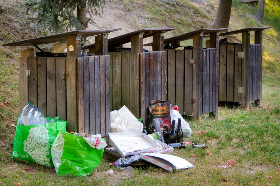 A row of four outdoor wooden rubbish bins with dark brown vertical planks and sloped roofs are situated on a grassy area near a woodland environment, with tall trees and a slope in the background. In front of the bins, there are various discarded items including a crumpled green plastic bag containing empty plastic bottles, a white pizza box with food remnants, a cardboard box labeled 'barbecue' partially crushed, and several glass bottles. Scattered around these objects are additional waste items such as plastic wrappers, and a few smaller bottles, showcasing an informal waste disposal scene. The setting appears to be an open space or park with natural light illuminating the scene, highlighting the textures and colors of the discarded waste and the weathered finish of the bin structures. This depiction relates to the topic of alternative waste handling or private rubbish collection, emphasizing the importance of proper rubbish disposal methods that waste management services like Rubbish Removal Holland Park can assist with.